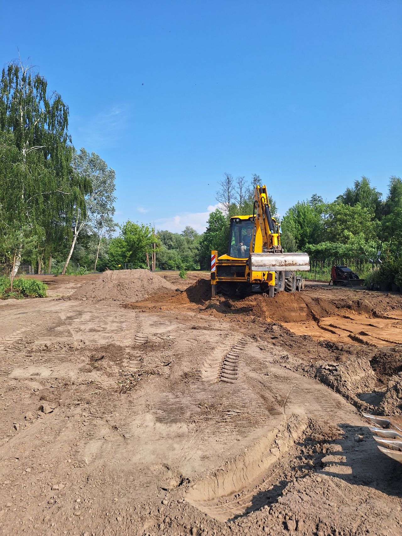 Kadr z prac ziemnych, niwelacji terenu. Żółta koparko-ładowarka zagarnia ziemię łyżką skarpową.